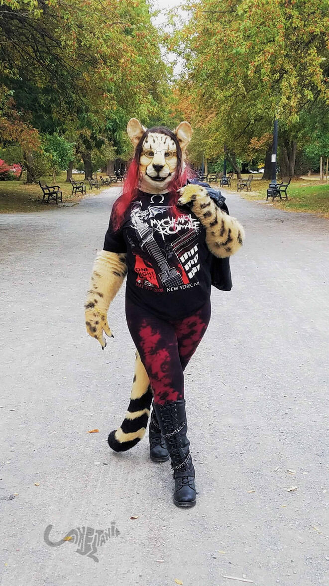 Shetani walking down a tree-lined park path wearing a MCR shirt, knee-high leather boots, leggings, and a leather jacket