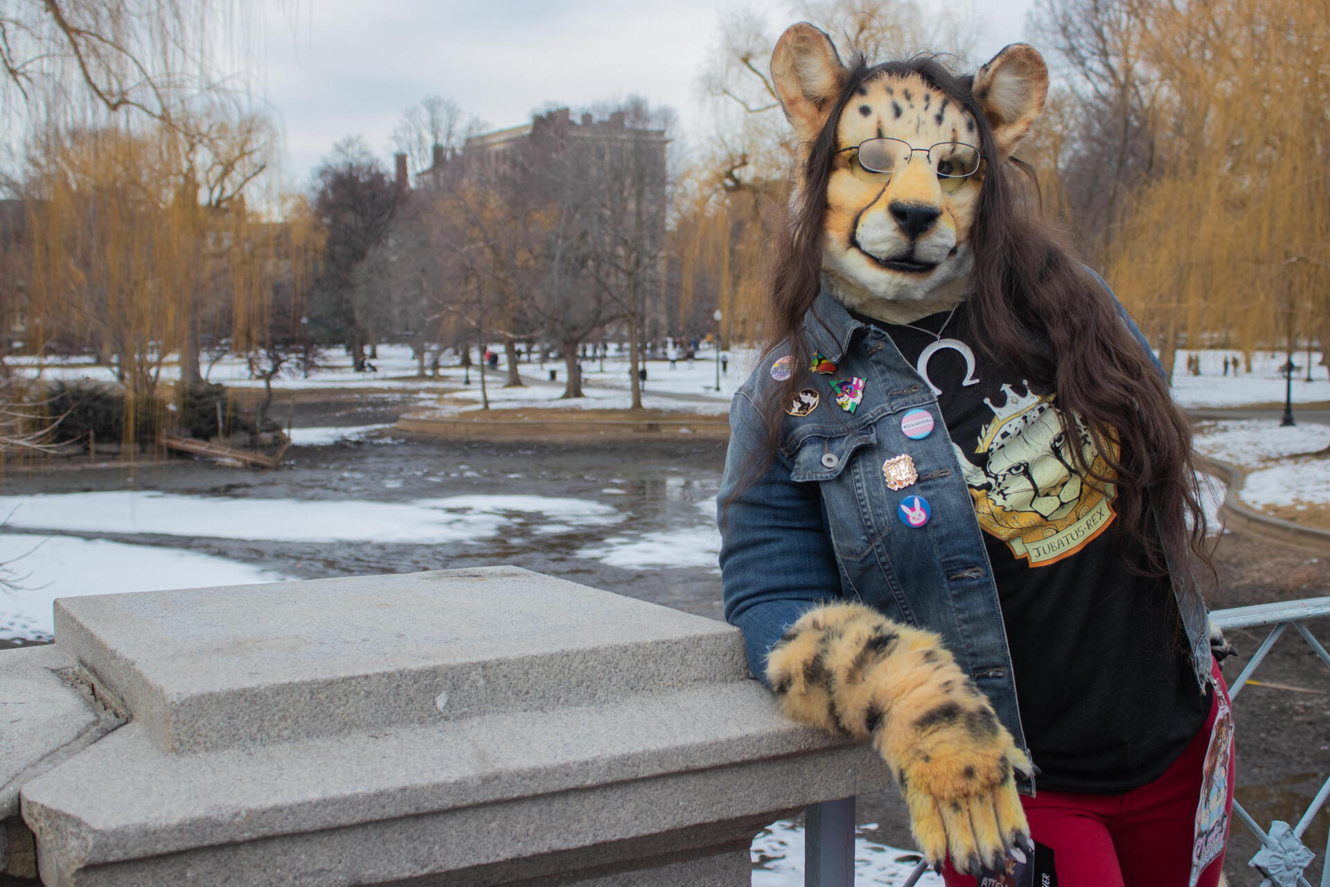 Shetani leaning on a bridge railing overlooking a frozen pond. Her breath fogs her glasses. Jacket front shows pins and buttons