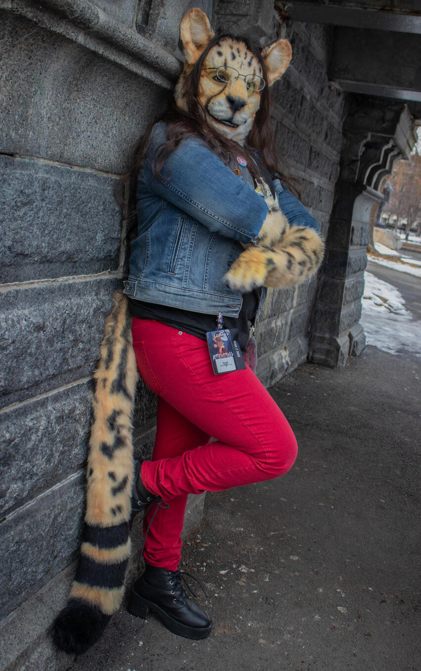 Shetani leaning against a wall, arms crossed and one foot up, wearing a denim jacket and red pants with boots