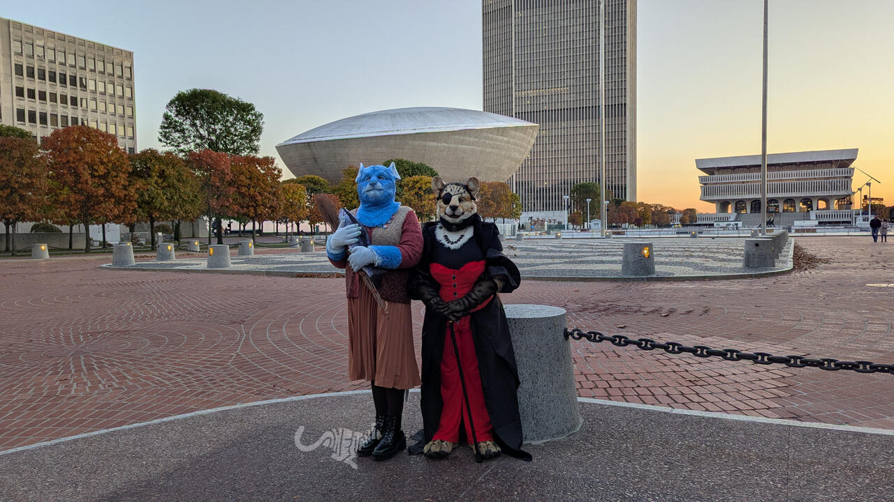 Two fursuiters in Halloween costume. Mika the blue rat is dressed as a poor humble citizen from a 1970s Soviet-inspired country, wearing a knit cardigan, skirt, and tights in brown hues, holding a fish and sheaf of wheat. Shetani the king cheetah is dresse