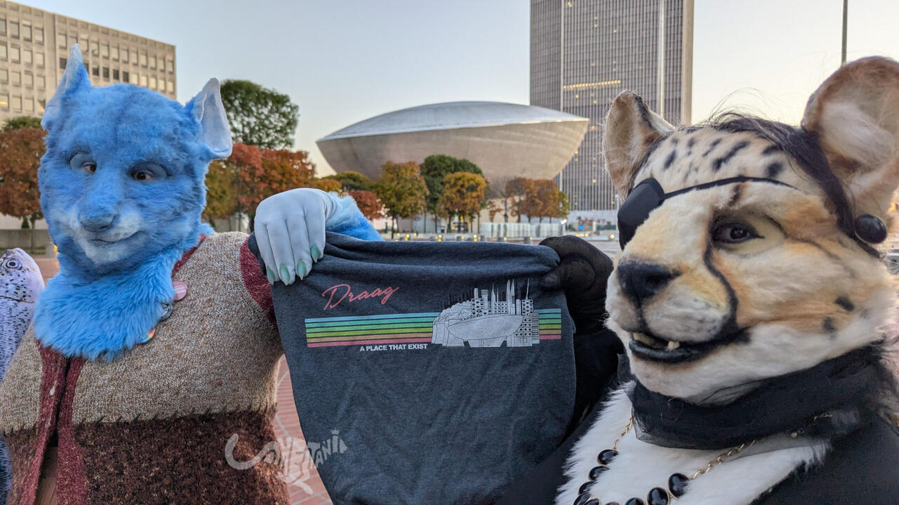 The two ladies are holding up a t-shirt with a graphic that reads "Draag - A Place That Exist" with a rainbow and some urban buildings. Prominently, the focal point of said buildings is the spitting image of The Egg they're standing in front of. Shetani is