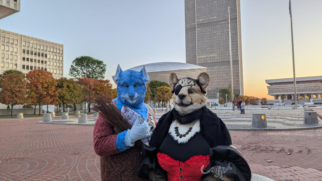 Waist-up shot of the pair in costume, allowing a clearer look at details such as Shetani's jeweled necklace, choker, and eye patch, and Mika's plush Gubrik fish and sheaf of wheat. Waist-up shot of the pair in costume, allowing a clearer look at details such as Shetani's jeweled necklace, choker, and eye patch, and Mika's plush Gubrik fish and sheaf of wheat.