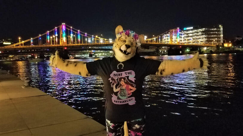 Shetani with arms spread wide in front of the illuminated bridge over the Anthrocon waterfront at nighttime