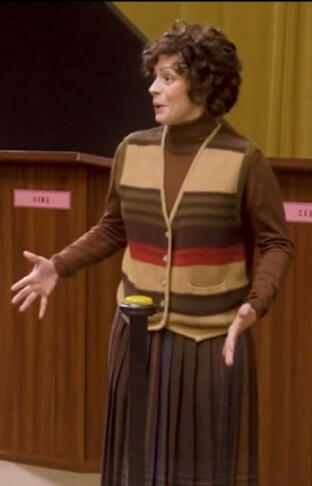Mika, a curly-haired woman wearing a 1970s-esque earth toned sweatervest, brown turtleneck sweater, and brown A-line skirt. Mika, a curly-haired woman wearing a 1970s-esque earth toned sweatervest, brown turtleneck sweater, and brown A-line skirt.