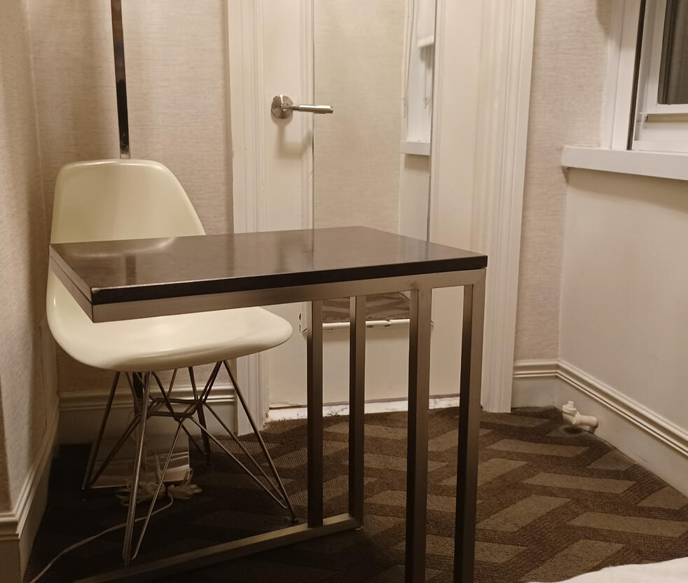 Hotel room from 2022, a plastic chair by a half desk on wheels. It smacks of cheap public school seating Hotel room from 2022, a plastic chair by a half desk on wheels. It smacks of cheap public school seating