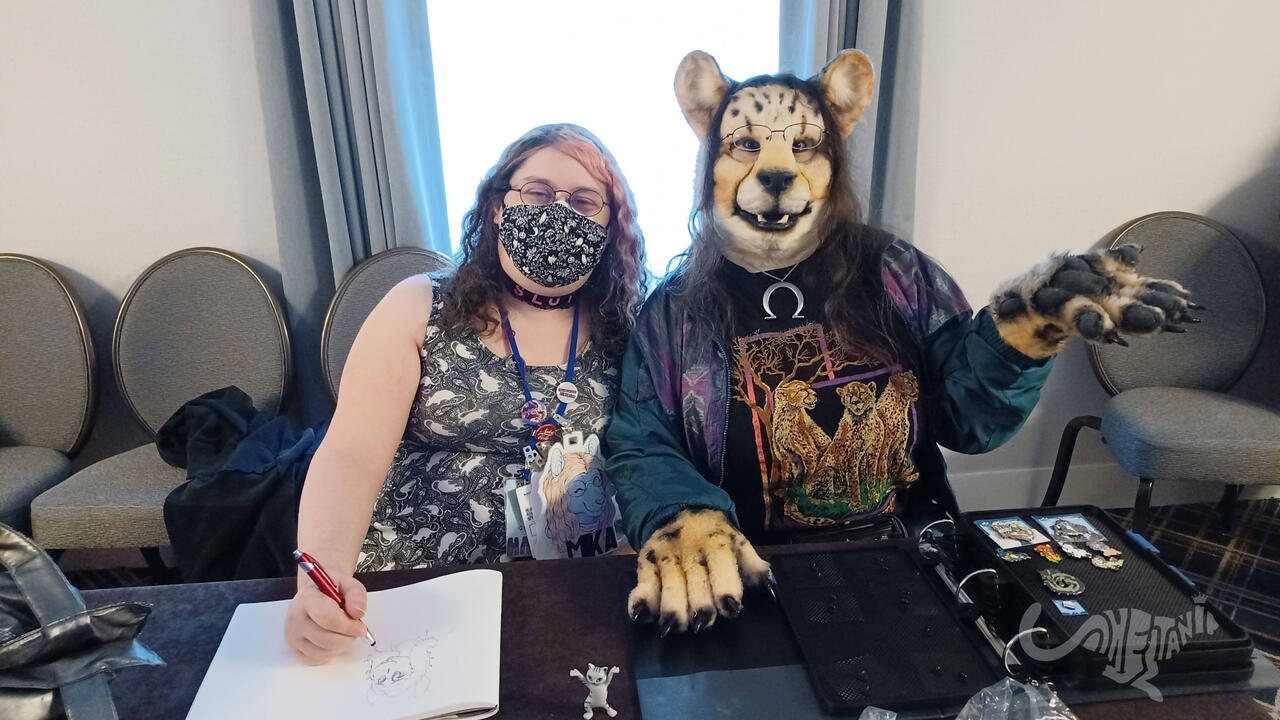 Mika seated at a table drawing in a sketchbook, with Shetani beside her gesturing to her open pin trading binder