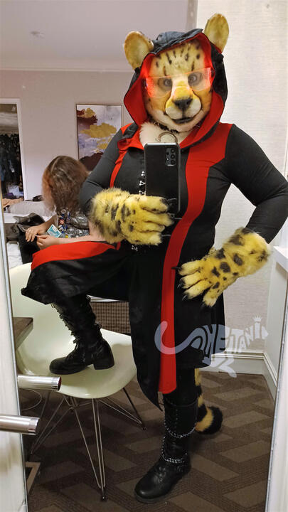 Mirror selfie of Shetani with one foot up to show her full outfit of a hooded black and red robe, black leggings, knee-high leather boots with chains, spiked collar, and glowing red cyberpunk visor. Mika is in background on the bed