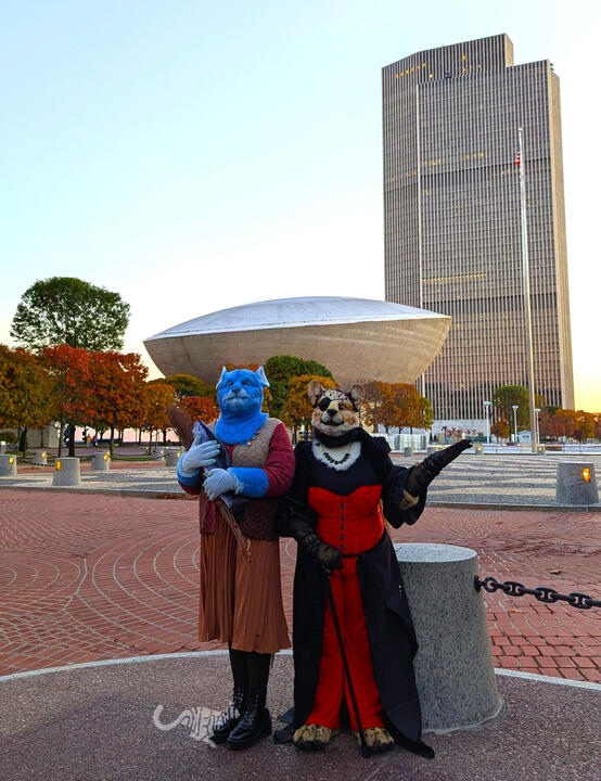 Full shot of the pair in costume posing beneath The Egg as the sun sets, Shetani gesturing with one arm grandly as timid Mika clings to her fish.