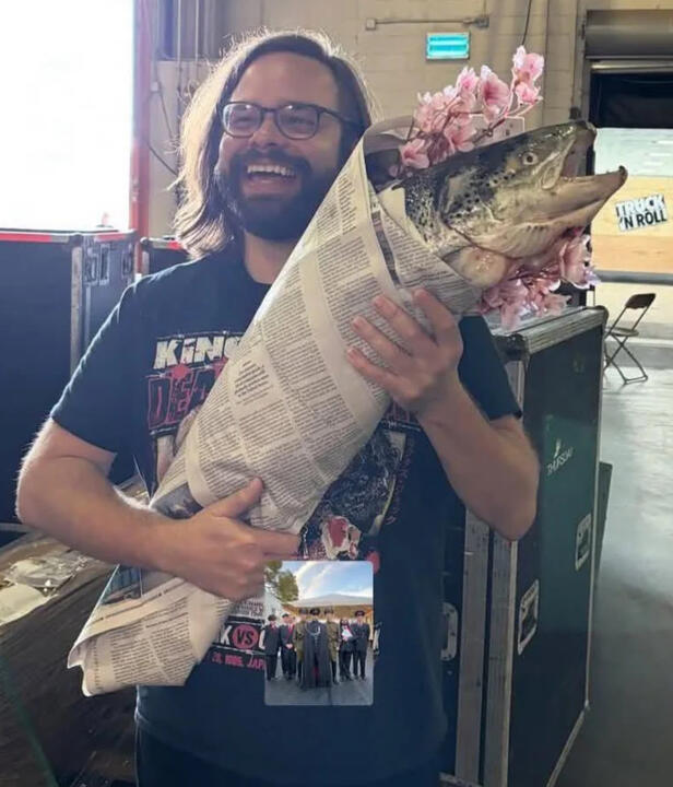 The actor who plays The Clerk backstage holding the fish, likely an Atlantic salmon, bundled in newspaper with cherry blossom branches.