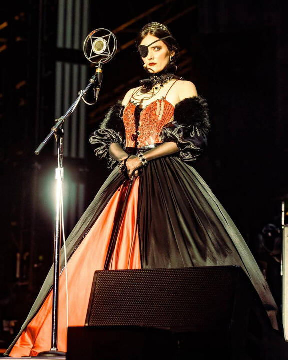 Full body photo of Sylvia on stage. She wears a red beaded corset, voluminous skirt with a black overskirt, black fur shawl, and uses a cane.