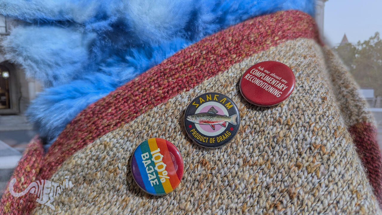 Three buttons pinned to Mika's earth-toned sweater. Top: white text on red "ask me about my complimentary reconditioning." Middle: "gubrik" in Keposhka and "product of Draag" in Latin alphabet surrounding a picture of a Gubrik fish. Bottom: "100% wheat" in