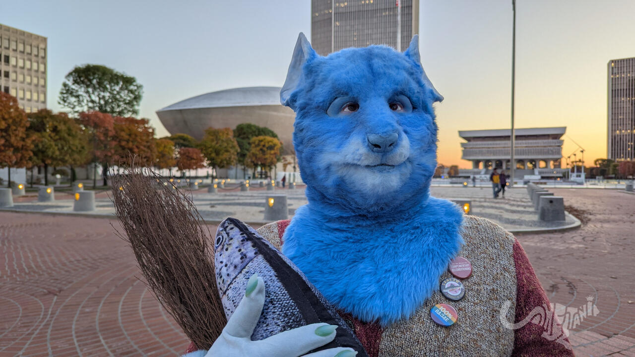 Mika the blue rat fursuiter standing at attention and gazing off into the distance. She's wearing a 1970s-esque cardigan in browns with three buttons pinned to it, and holding a sheaf of wheat and a fish. In the background, the sun sets behind a uniquely e
